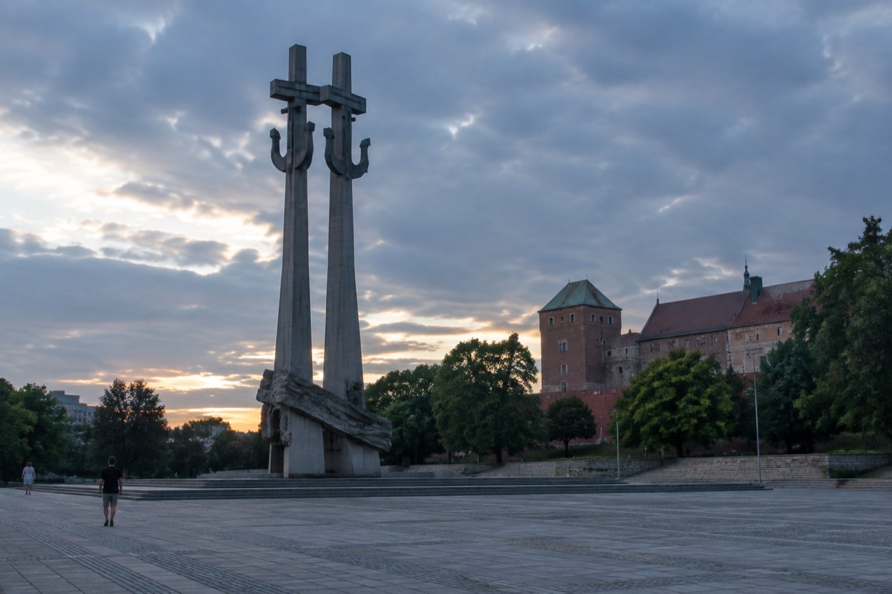 A tall stone monument with crossed sculpted forms standing on a wide plaza in central Poznan