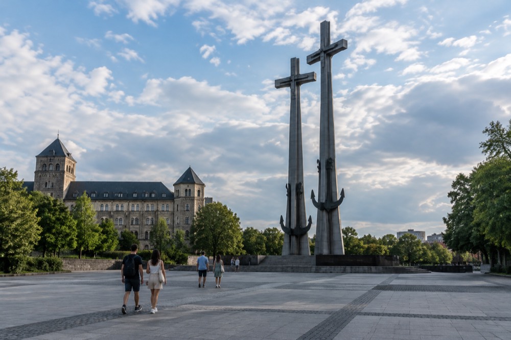 People walking across a city plaza toward a tall memorial sculpture on a sunny day in Poznan