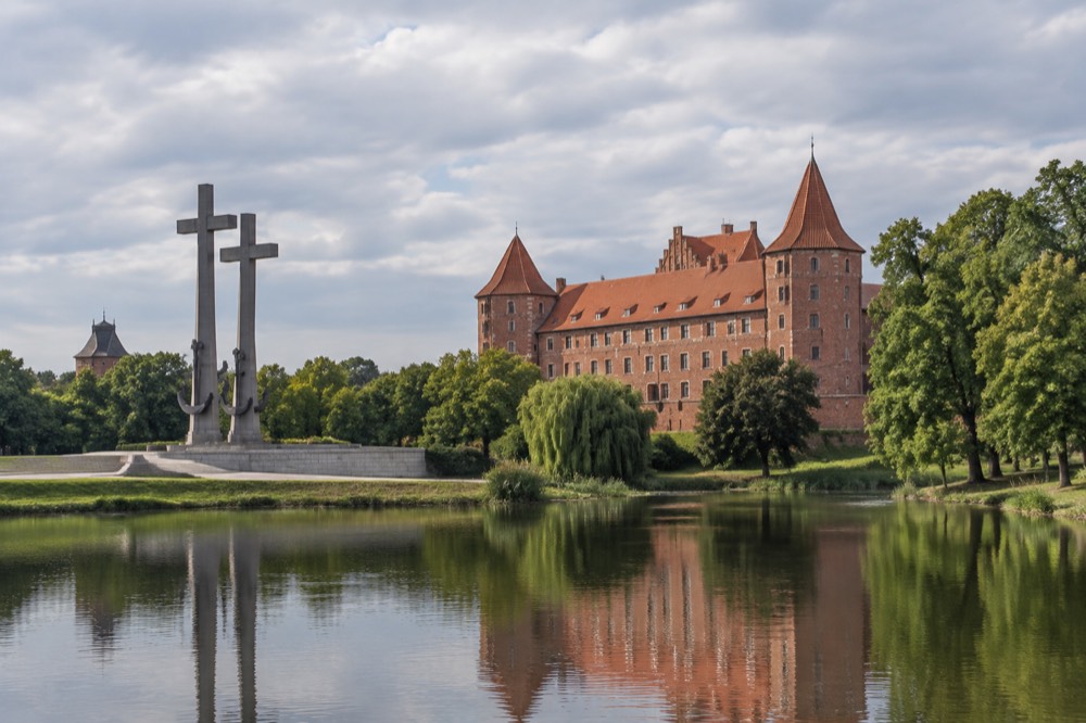 A red-brick castle reflected in water among large old trees at Kornik