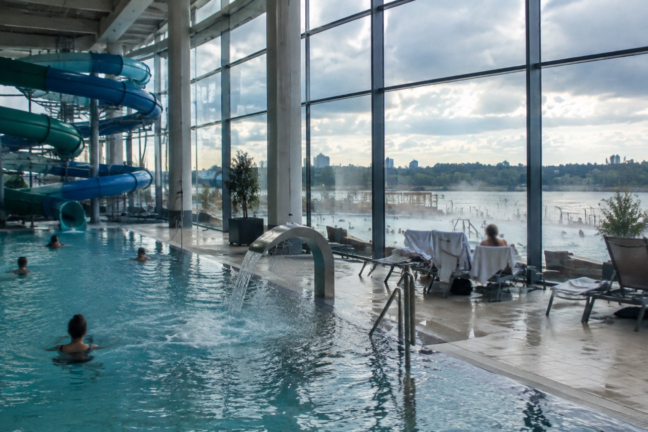 Indoor thermal pool hall with tall glass walls and water slides under daylight